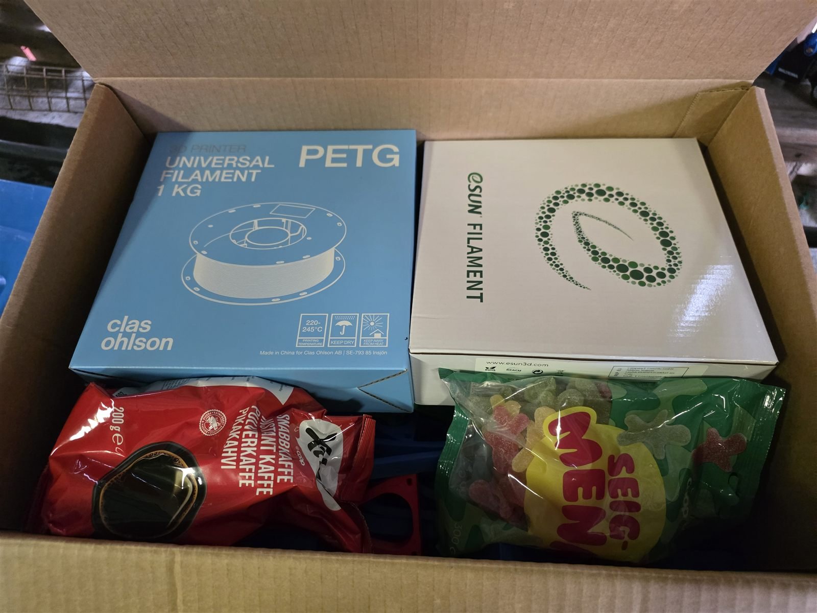 Åpen pappeske med to esker filament, kaffe og en pose seigmenn oppå de printede delene